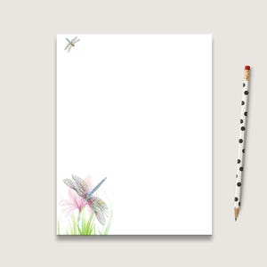 May include: White stationery paper with a watercolor illustration of a dragonfly and pink flowers. A black and white polka dot pencil rests on the right side of the paper. The dragonfly has blue and gray wings.