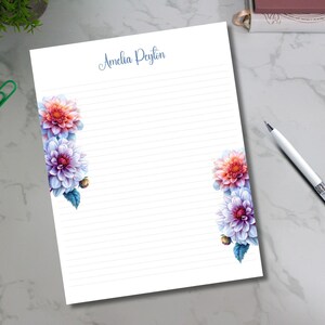May include: White lined stationery with floral accents. The stationery features watercolor-style dahlias in shades of red, purple, and blue. The name "Amelia Peyton" is written in script at the top. A pen rests on the side.