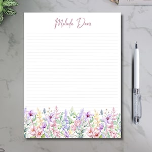 May include: A lined notepad with a floral border featuring pink, purple, and yellow flowers. The name "Melinda Davis" is written in cursive at the top. A silver and crystal pen rests on a marble surface.