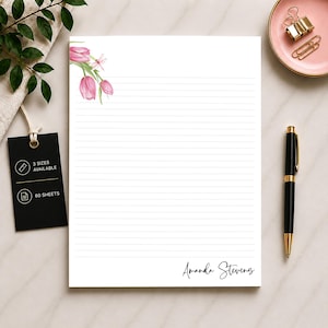 May include: White lined notepad with a pink floral design in the upper left corner. The notepad has the name "Amanda Stevens" printed in cursive at the bottom. A black pen, gold paper clips, and a pink dish are also visible.