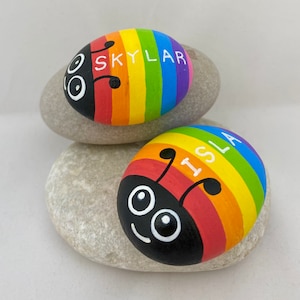 May include: Two painted rainbow ladybug rocks with the names "Skylar" and "Isla" written on them.