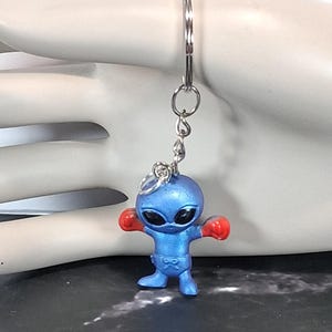 May include: A keychain featuring a small blue alien figurine wearing red boxing gloves. The alien has a smooth, rounded head with large, dark, oval-shaped eyes and a compact body. It is attached to a silver-toned metal split ring by a short chain.