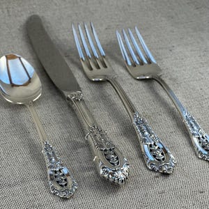 May include: A set of four silver antique cutlery items: a spoon, a knife, and two forks. The handles have elaborate floral and scrollwork patterns. The cutlery is arranged on a neutral-coloured linen surface.