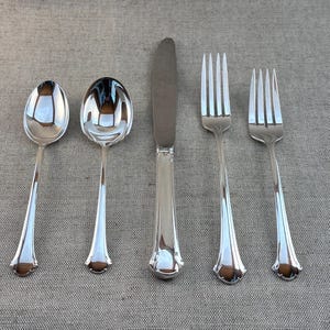 Puede incluir: Un juego de cinco cubiertos pulidos de color plateado, que incluye dos cucharas, un cuchillo y dos tenedores. Los utensilios están dispuestos sobre una superficie de color neutro, mostrando sus cualidades reflectantes y su elegante diseño.