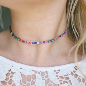 Peut inclure: Un collier ras de cou composé de petites perles multicolores. Le collier est fait d'une fine chaîne et est porté par une personne aux cheveux blonds.