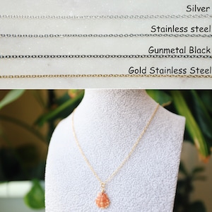 May include: A gold stainless steel necklace with a small, orange seashell pendant. The necklace is displayed on a white bust, with a blurred green background. The image also shows examples of silver, stainless steel, and gunmetal black chain.