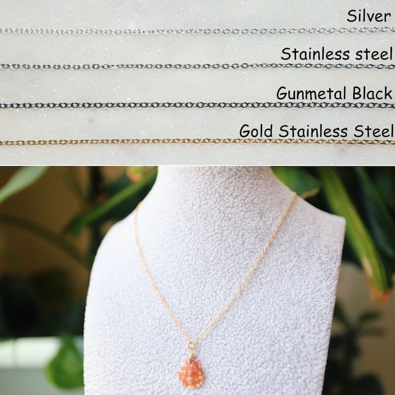May include: A gold stainless steel necklace with a small, orange seashell pendant. The necklace is displayed on a white bust, with a blurred green background. The image also shows examples of silver, stainless steel, and gunmetal black chain.