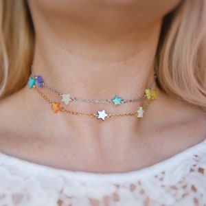 May include: A delicate layered necklace with a silver chain and a gold chain. Both chains are adorned with small star-shaped charms in various pastel colors, including turquoise, purple, yellow, and orange.