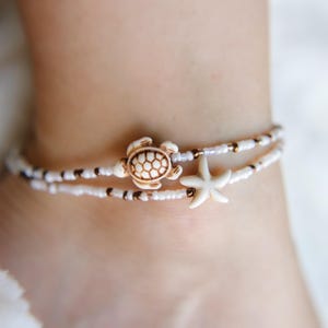 May include: Ankle bracelet featuring two strands of white beads with brown accents. The top strand has a turtle charm, and the bottom strand has a starfish charm. The bracelet is worn on an ankle.