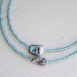 May include: Two delicate necklaces with light blue seed beads. Each necklace features a rectangular, iridescent abalone shell pendant. The necklaces are displayed on a white surface, creating a contrast with the blue beads and pendants.