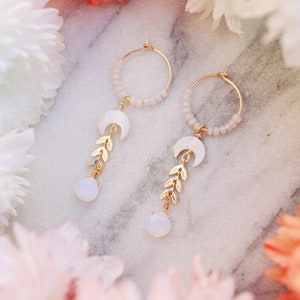 May include: A pair of gold hoop earrings with delicate white beads and a crescent moon charm with gold leaf accents hanging from each hoop. The earrings are accented with white opal stones.