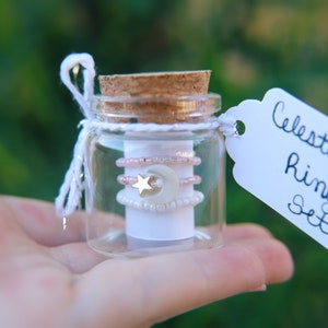 May include: A small glass jar with a cork lid contains a set of delicate rings with a star and crescent moon charm. The jar has a white tag attached that reads "Celestial Ring Set".