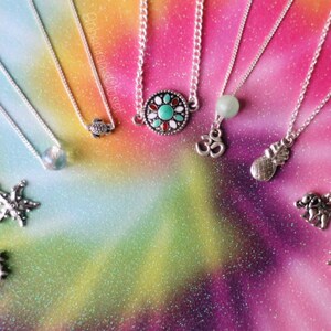 Pu&ograve; includere: Dieci collane d'argento con ciondoli diversi, tra cui una tartaruga, una stella marina, una stella, un mandala turchese e argento, una perla verde, un simbolo Om, un ananas, un elefante e un sole. Le collane sono esposte su uno sfondo tie-dye colorato.