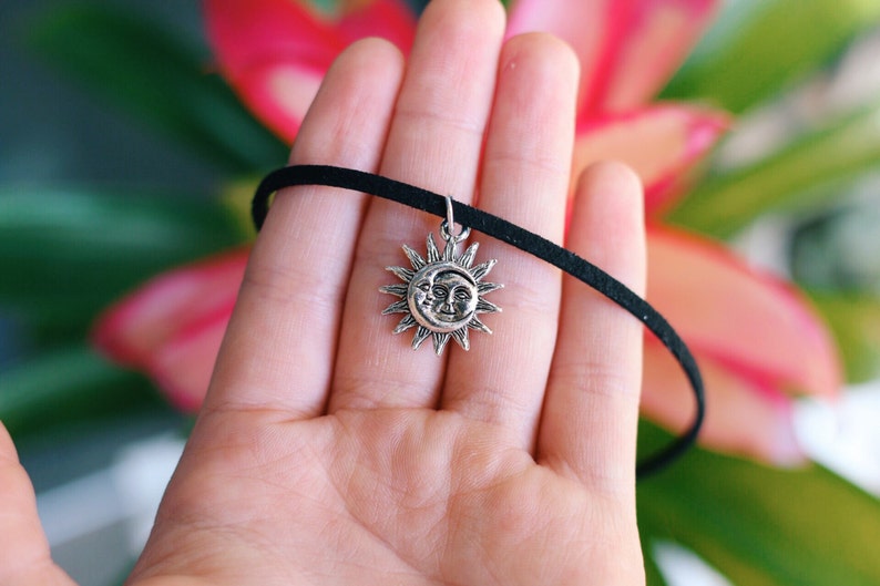 May include: A black choker necklace with a silver sun and moon pendant. The pendant features a detailed design of a sun with a crescent moon inside.
