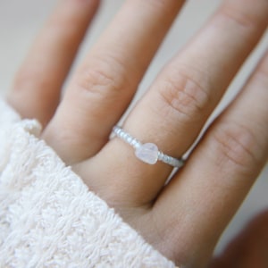 May include: A delicate ring with a small, heart-shaped, white gemstone set in a silver beaded band.