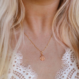 May include: A gold-colored necklace with a small, ornate pendant. The pendant has a cluster of small, light-colored stones. The necklace is worn against a background of blonde hair and a white lace top.
