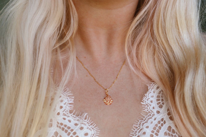 May include: A gold-colored necklace with a small, ornate pendant. The pendant has a cluster of small, light-colored stones. The necklace is worn against a background of blonde hair and a white lace top.