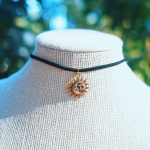 May include: A black choker necklace with a gold sun and moon pendant. The pendant features a stylized sun with a crescent moon inside.
