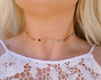 May include: A delicate gold-tone choker necklace adorned with a series of colorful gemstones. The necklace features a variety of jewel tones, including red, orange, yellow, green, and blue. The necklace is worn against a white lace top.