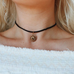 May include: A black choker necklace with a gold sun pendant. The sun has a face and rays.