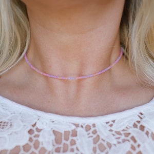 May include: A delicate choker necklace made with small, colorful beads in shades of pink, purple, and white. The necklace is worn by a person with blonde hair and a white lace top.