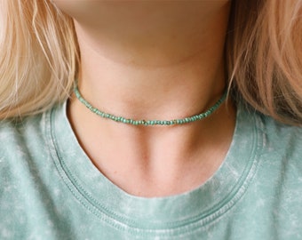 May include: A delicate choker necklace composed of small, turquoise-colored beads. The necklace sits around the neck, creating a simple and elegant accessory. The beads are evenly spaced, forming a continuous strand. The necklace is a light, bright turquoise color.