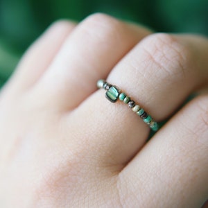 May include: A delicate beaded ring with a turquoise and brown color scheme. The ring features a larger, rectangular turquoise bead in the center.
