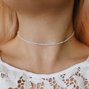 May include: A delicate white beaded choker necklace worn on a model's neck. The necklace is made of small, evenly spaced beads and is a simple, minimalist design.