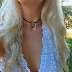 May include: A woman with long blonde hair wearing a white lace top with a black choker necklace featuring a gold sun pendant.