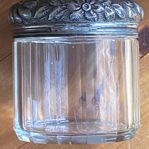 May include: A clear glass jar with vertical ridges and a decorative silver lid. The lid features a floral design around the edge. The jar is cylindrical and sits on a wooden surface. This antique jar could be used for storage.