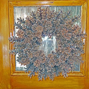 Op de afbeelding: Een bruine dennenappelenkrans met geglazuurde dennenappelen hangt aan een houten deur.