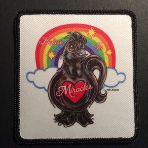 Puede incluir: Un parche bordado en blanco y negro con un arcoíris, nubes y un pájaro negro con un corazón rojo que dice "Miracles" en su interior. El texto "Believe in" está por encima del pájaro.