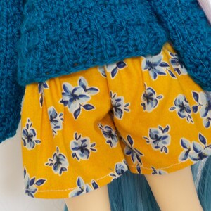 May include: A doll wearing a teal blue knit sweater and yellow floral shorts. The doll has long, white hair with pink highlights and blue hair extensions. The doll's eyes are closed, and it is set against a white background.