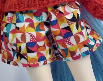 May include: A doll wearing a red knitted sweater and colorful patterned shorts. The shorts feature a geometric design with shapes in yellow, pink, blue, and black. The doll has light skin and blue hair.
