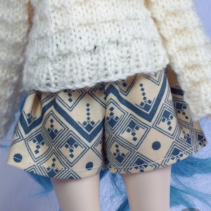 May include: A doll wearing a white crocheted sweater and a yellow and blue patterned skirt.