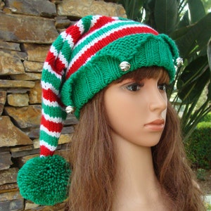 Peut inclure: Un bonnet en tricot rayé vert, rouge et blanc avec un long pompon vert et des grelots. Le bonnet est porté sur un mannequin aux longs cheveux bruns.