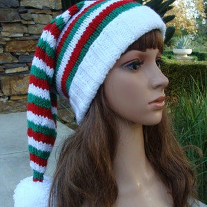 May include: A white, red, and green striped knit hat with a long, pointed top and a white pom-pom. The hat is worn on a mannequin with long brown hair.