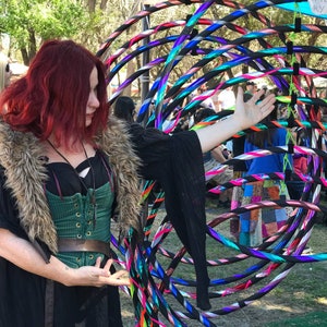 May include: A woman with long red hair wearing a black shirt with a fur collar and a green corset is holding a hoop. The hoop is made of multiple colorful plastic rings. The woman is standing in a field with other people in the background.
