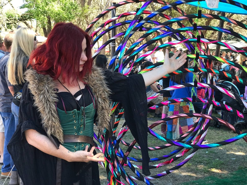 May include: A woman with long red hair wearing a black shirt with a fur collar and a green corset is holding a hoop. The hoop is made of multiple colorful plastic rings. The woman is standing in a field with other people in the background.
