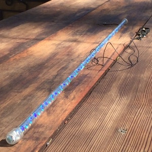 Może przedstawiać: Przezroczysta, cylindryczna r&oacute;żdżka wypełniona iryzującym, niebieskim i fioletowym brokatem. R&oacute;żdżka ma przezroczystą, zaokrągloną końc&oacute;wkę i ozdobny metalowy element z drutami. R&oacute;żdżka spoczywa na drewnianej powierzchni.