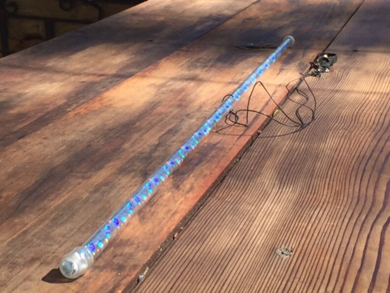 Może przedstawiać: Przezroczysta, cylindryczna r&oacute;żdżka wypełniona iryzującym, niebieskim i fioletowym brokatem. R&oacute;żdżka ma przezroczystą, zaokrągloną końc&oacute;wkę i ozdobny metalowy element z drutami. R&oacute;żdżka spoczywa na drewnianej powierzchni.