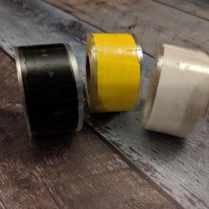 May include: Three rolls of tape, one black, one yellow, and one white. The rolls are on a wooden surface.