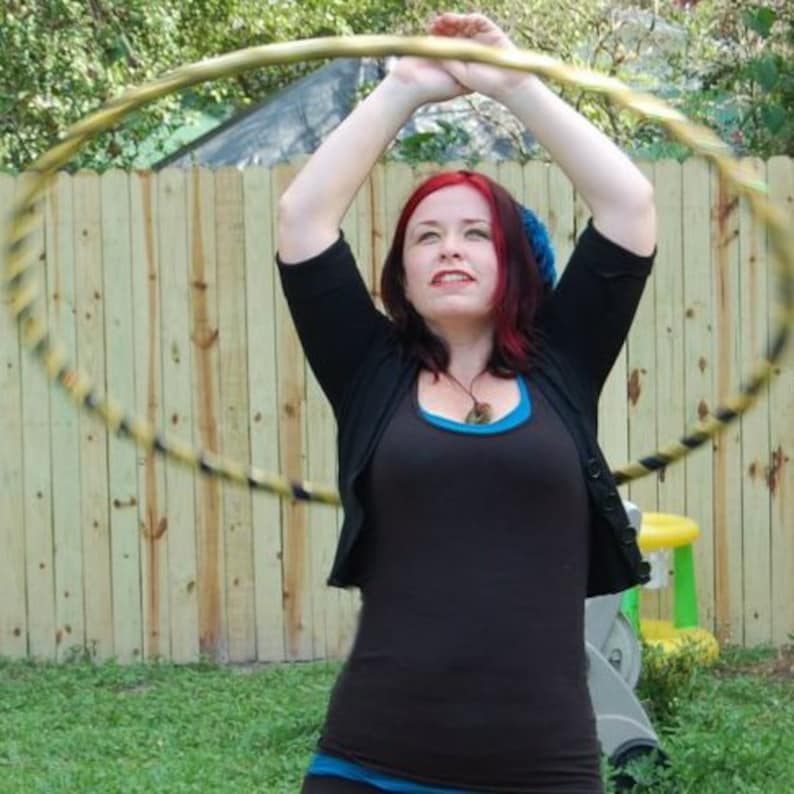 May include: A woman with red hair is holding a wooden hoop with black tape around it. She is wearing a black cardigan and a blue tank top. The hoop is in front of a wooden fence.