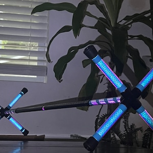 Può includere: Un paio di bastoncini luminosi blu e viola collegati da un'asta nera. I bastoncini luminosi hanno un motivo geometrico e sono incrociati a forma di X.