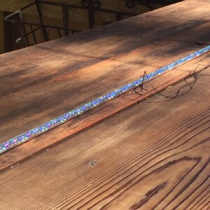 Może przedstawiać: Przezroczysta, cylindryczna r&oacute;żdżka wypełniona iryzującym brokatem. R&oacute;żdżka ma około 61 cm długości i ma przezroczystą nasadkę na jednym końcu. Do r&oacute;żdżki przymocowany jest mały, czarny element z przewodami.