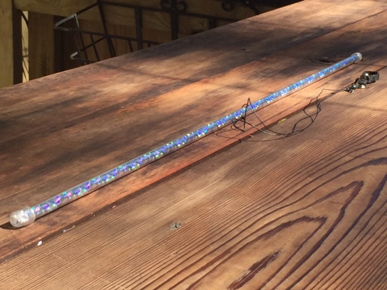 Może przedstawiać: Przezroczysta, cylindryczna r&oacute;żdżka wypełniona iryzującym brokatem. R&oacute;żdżka ma około 61 cm długości i ma przezroczystą nasadkę na jednym końcu. Do r&oacute;żdżki przymocowany jest mały, czarny element z przewodami.