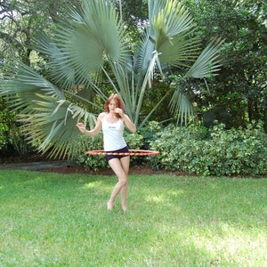 May include: A woman in a white tank top and black shorts is hula hooping in a grassy area. She is standing in front of a large palm tree.