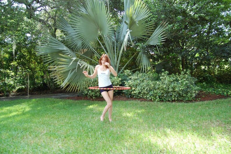 May include: A woman in a white tank top and black shorts is hula hooping in a grassy area. She is standing in front of a large palm tree.