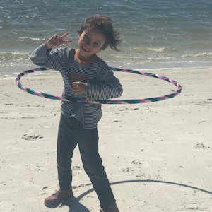 May include: A child is smiling and holding a colorful hula hoop on a white sandy beach. The hula hoop has pink, blue, black, and white stripes.
