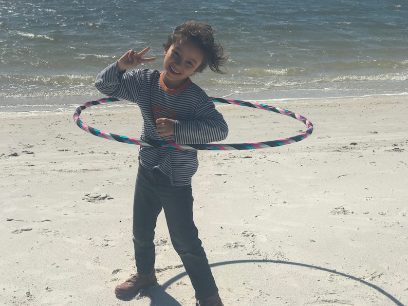May include: A child is smiling and holding a colorful hula hoop on a white sandy beach. The hula hoop has pink, blue, black, and white stripes.